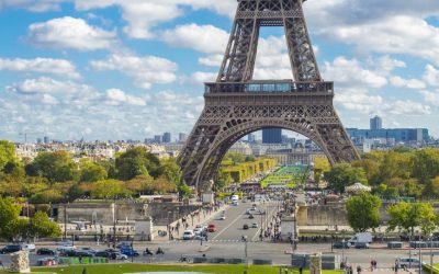 A close-up view of the Eiffel Tower's intricate wrought-iron lattice structure in Paris, France. The tower's curved arches and diagonal braces create a beautiful geometric pattern against a bright blue sky. A small group of tourists can be seen ascending the tower's stairs to reach the summit