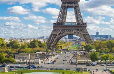 A close-up view of the Eiffel Tower's intricate wrought-iron lattice structure in Paris, France. The tower's curved arches and diagonal braces create a beautiful geometric pattern against a bright blue sky. A small group of tourists can be seen ascending the tower's stairs to reach the summit