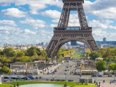 A close-up view of the Eiffel Tower's intricate wrought-iron lattice structure in Paris, France. The tower's curved arches and diagonal braces create a beautiful geometric pattern against a bright blue sky. A small group of tourists can be seen ascending the tower's stairs to reach the summit