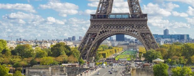 A close-up view of the Eiffel Tower's intricate wrought-iron lattice structure in Paris, France. The tower's curved arches and diagonal braces create a beautiful geometric pattern against a bright blue sky. A small group of tourists can be seen ascending the tower's stairs to reach the summit