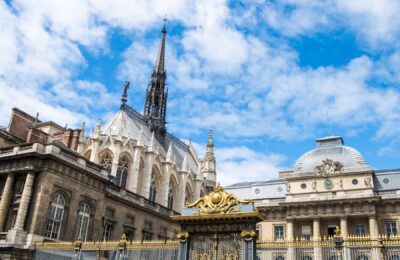 A photo of the exterior of Sainte Chapelle in Paris, a Gothic-style church with two tiers of intricately detailed stained-glass windows and pointed arches, situated on a cobblestone street with a tree-lined background.