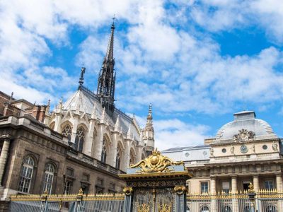 A photo of the exterior of Sainte Chapelle in Paris, a Gothic-style church with two tiers of intricately detailed stained-glass windows and pointed arches, situated on a cobblestone street with a tree-lined background.